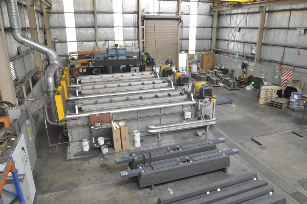 A large industrial warehouse with several long metal beams and machines lined up. The space is filled with equipment and supplies, with a few workers visible in the background. The walls are made of sheet metal with tall ceilings.