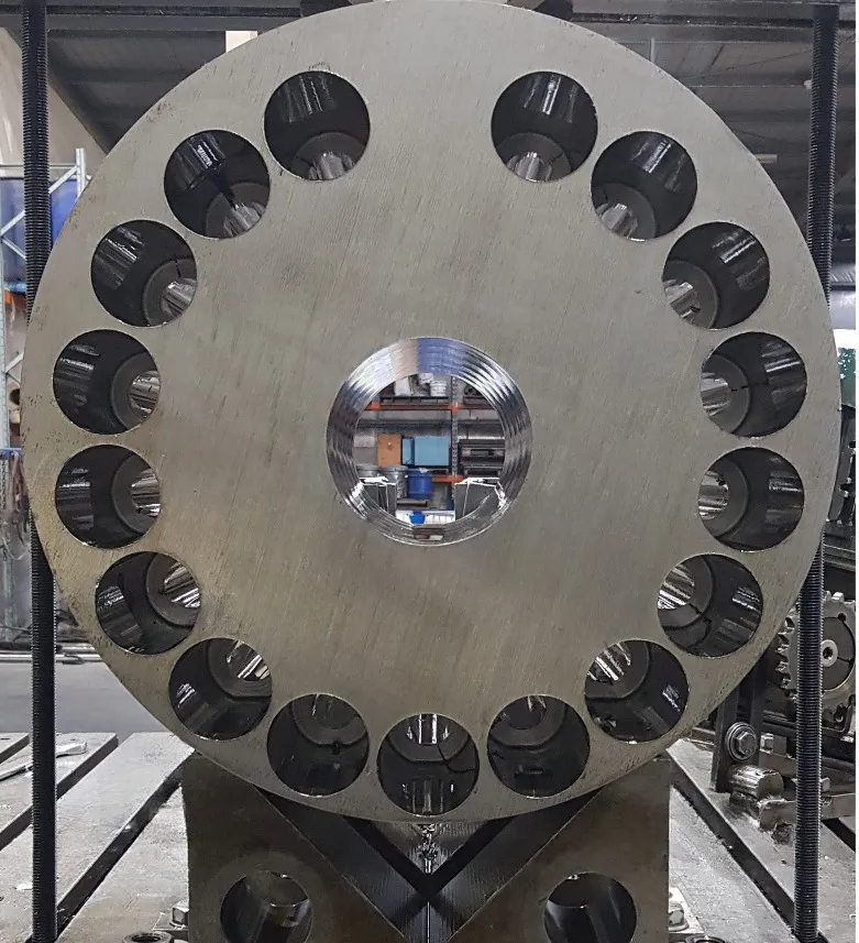 A large circular metal object with multiple evenly spaced circular holes around its perimeter. The object is positioned vertically and appears to be part of a mechanical or industrial setup in a workshop or factory.