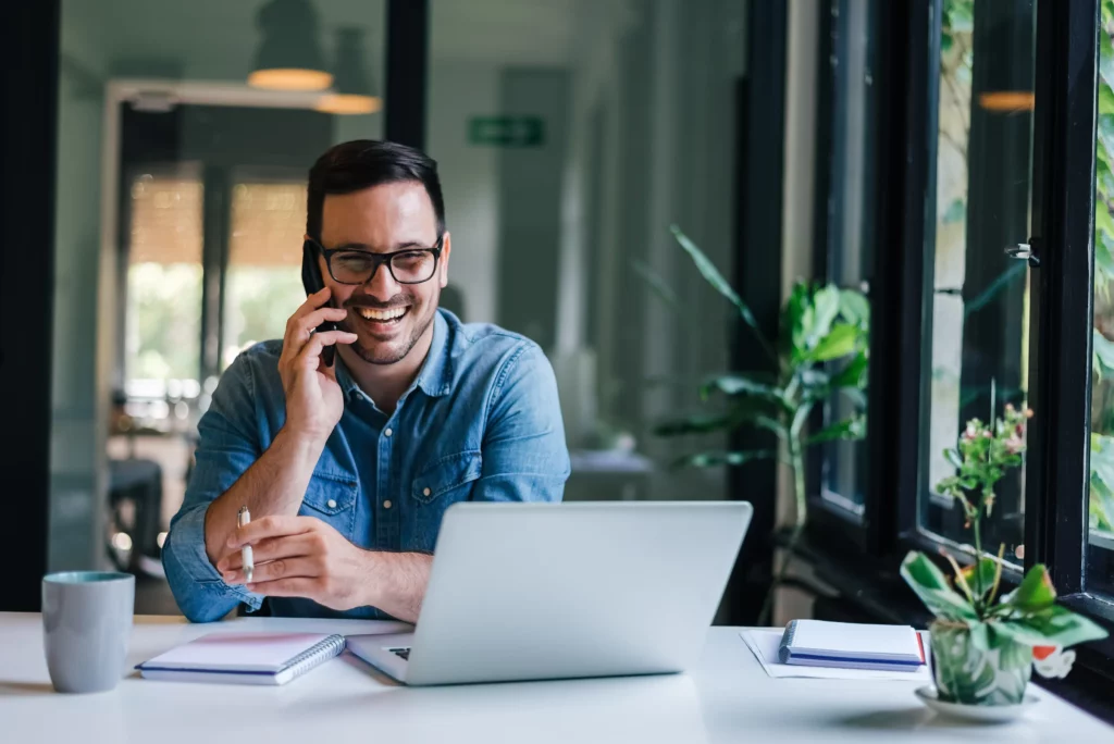 A smiling man wearing glasses and a denim shirt is talking on the phone while sitting at a desk with a laptop and notebooks. There's a mug on the desk and a window with plants in the background.