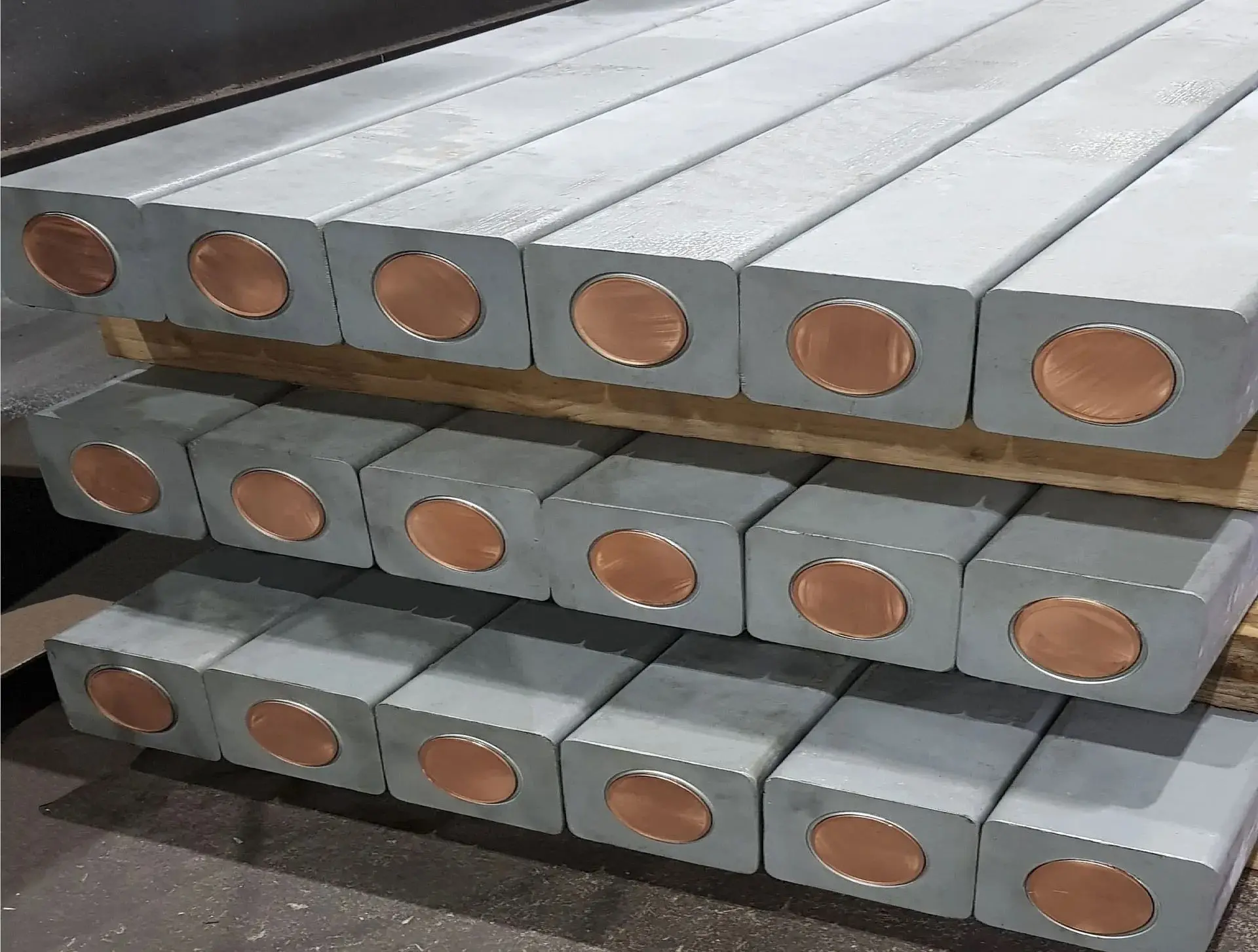 Stacks of rectangular metal bars with circular copper ends are arranged neatly on wooden pallets in an industrial setting.