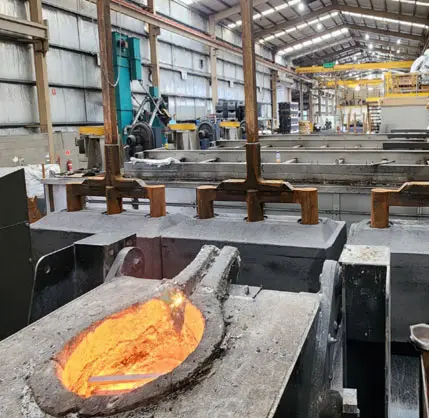 A foundry interior with molten metal glowing orange in a crucible, large metal molds, overhead cranes, and industrial equipment visible inside a spacious, high-ceilinged warehouse.