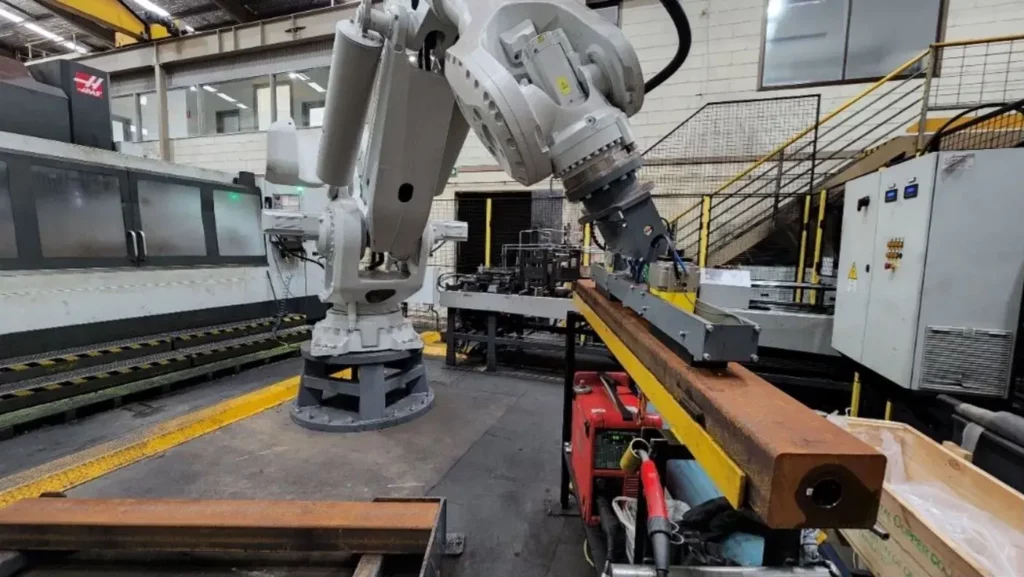 A large industrial robotic arm works on a long metal beam in a spacious factory setting, surrounded by various machinery and metal structures with stairs and railings in the background.