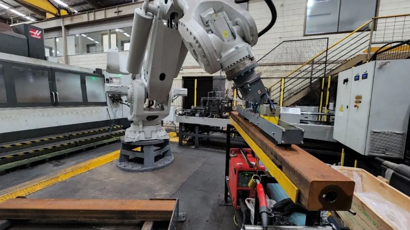 A large industrial robotic arm works on a long metal beam in a spacious factory setting, surrounded by various machinery and metal structures with stairs and railings in the background.
