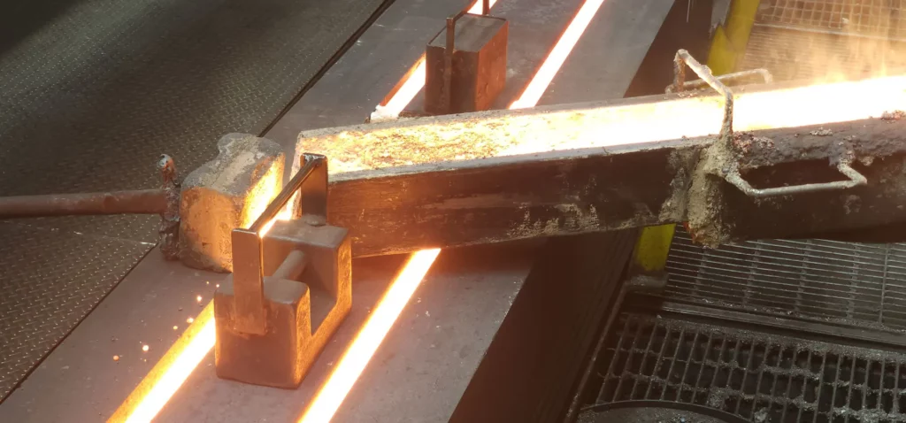Molten metal is being poured from a tilted rectangular mold, glowing orange from the heat, onto a metal surface in an industrial setting. The surrounding area includes metal grates and safety barriers.