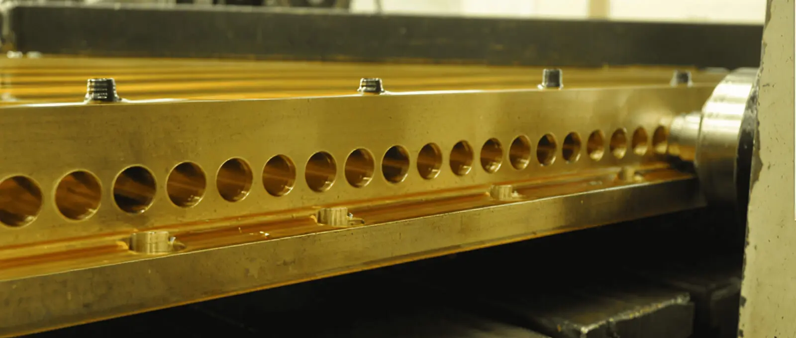 Close-up of a metal piece with evenly spaced circular holes being machined, with a cutting tool visible on the right side. The metal appears to be mounted on industrial equipment.
